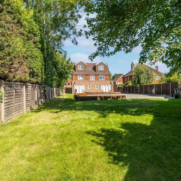 A view from the far end of the garden looking back at the building, with a lawn area under a tree and a parking area in the middle ground.