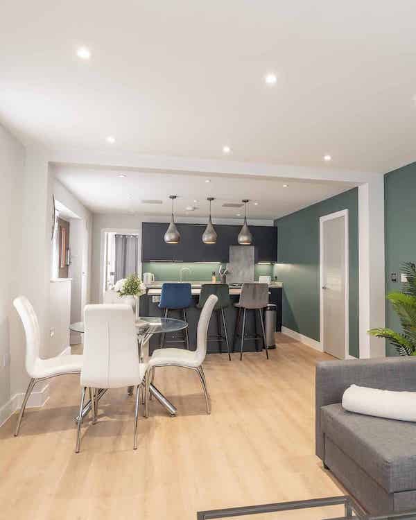 A modern living area showing four chairs around a glass round table and in the background a kitchen area with three bar stools facing a breakfast bar