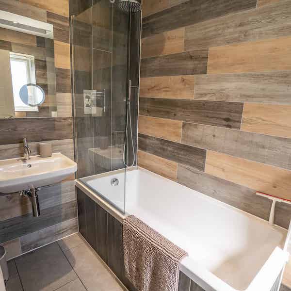 A bath with integrated shower and a glass panel. On the left of the image is a sink below a mirror.